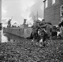 British soldiers assist a wounded soldier between buildings towards the sea and a waiting landing craft left