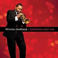 Before a red background, a man in a suit is playing a trumpet with his eyes closed; the text "Wynton Marsalis Christmas Jazz Jam" appears in a black bar across the center