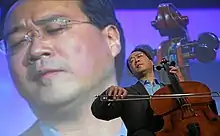 A man playing on a cello, with his eyes closed and his body visible on a large screen