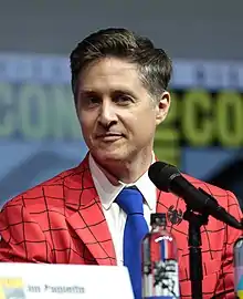 A middle aged caucasian male looks slightly to the leftwith a small smile. He has short brown hair and is wearing a Spider-Man-themed red coat with webbing trim