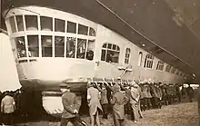 The gondola while the airship is being manoeuvred on the round. Around forty people on the ground are manhandling it. Two officers are visible in the gondola, one looking down at the people, the other looking backwards. The ram air turbine is folded flush with the gondola's side.