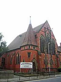 A red brick chapel with stone dressings with a small spire in the foreground and a noticeboard announcing "Zion Tabernacle"