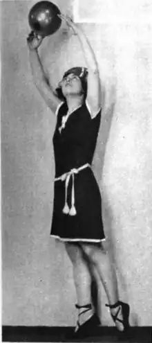 A young white woman, arms overhead as if to catch the ball just beyond her fingertips. She is wearing a dark jersey one-piece dress-like bathingsuit with white piping and a belt with tassles. She is also wearing ballet-style slippers, and a cap.