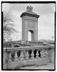 A stone arch with stone eagle is in the background near the entrance to a bridge that continues beyond it. A stone banister and railing in the foreground.