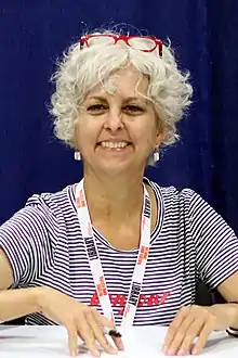 Kate DiCamillo at the 2018 U.S. National Book Festival smiling at the camera holding a pen with red glasses resting on top of her head.