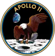 Circular insignia: eagle with wings outstretched holds olive branch on Moon with Earth in background, in blue and gold border.