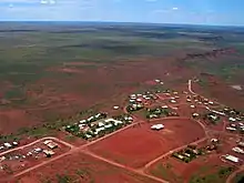 An aerial view of a small settlement, showing about seventy rooftops, red dirt roads and a dirt oval, with dry scrubland receding into the distance