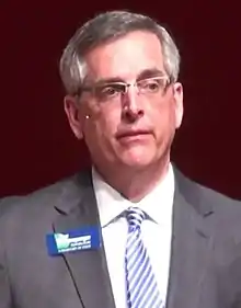 Brad Raffensperger wearing a grey suit with a name tag.