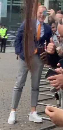 Full length view of a tall young man standing next to security barriers lined with fans some having cameras ready