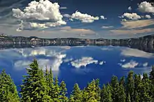 Flat, blue lake clearly reflecting a cloudy sky, fronted by pine forest