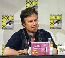 A Caucasian man in his forties, seated at a conference, with a microphone in front of him. He has a square face, deep-set eyes, dark hair and a brown beard with clean-shaved cheeks and upper-lip. He is casually dressed, relaxed and smiling. Square signs are posted on the wall behind him, bearing the name COMIC-CON in big bright yellow letters around a drawn eye and eyebrow.