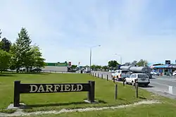 A brown sign on the side of the road says "Darfield". Several cars are parked on the side of the road. Trees are visible to the left and buildings in the distance.