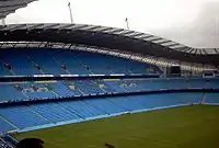 Roughly the same camera position shows grass up to the blue seats of the stands. The stand is now split into three levels of permanent seating.