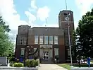 A Moderne-style brick and stone rectangular courthouse structure with a clock tower and flagpole on the right side. The large rectangular windows of the second level reflect a bright blue sky with large white clouds