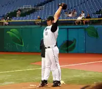 A man stretches his arms behind his head while wearing a baseball glove and a white baseball uniform with green sleeves and cap.