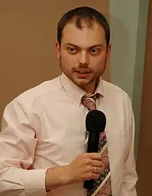 A standing bearded Russian man while wearing a pink shirt and striped necktie while holding a wireless black microphone; on his right index finger is a shiny golden wedding ring.