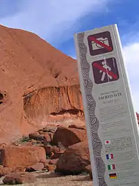 Tall sign with no entry and no photos symbols, in front of the base of a tall red rock