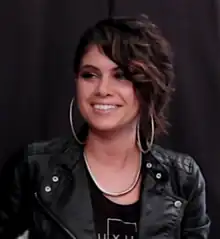 Still image of a woman looking away from the camera and smiling with an undercut hairstyle; wearing large hoop earrings, a black top, a thick necklace and a leather jacket.