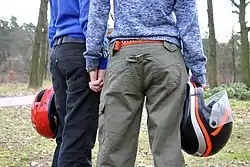 Photo of two women from the back from the shoulders down, dressed in outdoor clothing, carrying motorcycle helmets, and holding hands