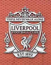 The words "Liverpool Football Club" are in the centre of a pennant, with flames either side. The words "You'll Never Walk Alone" adorn the top of the emblem in a green design, "EST 1892" is at the bottom