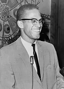 A 38-year-old man in a suit and tie smiles broadly. He wears glasses and has a microphone around his neck.