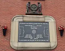 A stone tablet, inscribed with the image of a football pitch and several names. It is surrounded by a stone border in the shape of a football stadium. Above the tablet is a wooden carving of two men holding a large wreath.