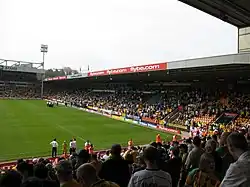 The inside of an association football stadium, with a stand on the right-hand side full of supporters. The pitch is visible to the left of the stand, with a floodlight in the background.