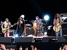 The Red Hot Chili Peppers performing on stage in 2006. From left to right: Flea is playing a bass guitar, Anthony Kiedis is singing, Chad Smith is playing a set of drums and John Frusciante is playing a guitar.