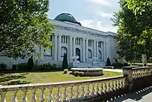 A white stone library with a green dome on top and two lions in front surrounded by grass and bushes