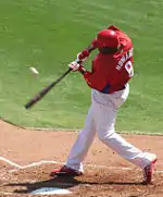 A man in white pants and a red shirt with "HOWARD" on the back swings at a pitch.