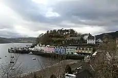 A small harbour fronted with a row of cottages painted in white, pink, green and blue with a tree-covered hillock behind them.