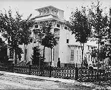 A small, white house with a man in a tuxedo and top hat looking at the house