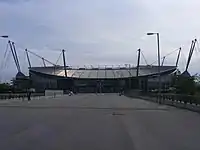 A straight tarmac road. At the head of the road is a stadium with a bowl-shaped outline, surrounded by a number of masts, with cables running to the stadium roof