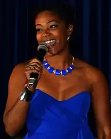 A Black woman in a blue dress in front of dark blue curtains