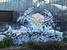 Wildstyle graffiti done on a corrogated iron wall, with a large crab character in the background