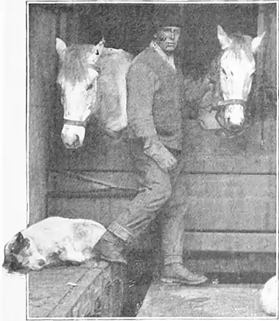 Photo of a man standing in front of two narrow horse stalls, with a sleeping dog him