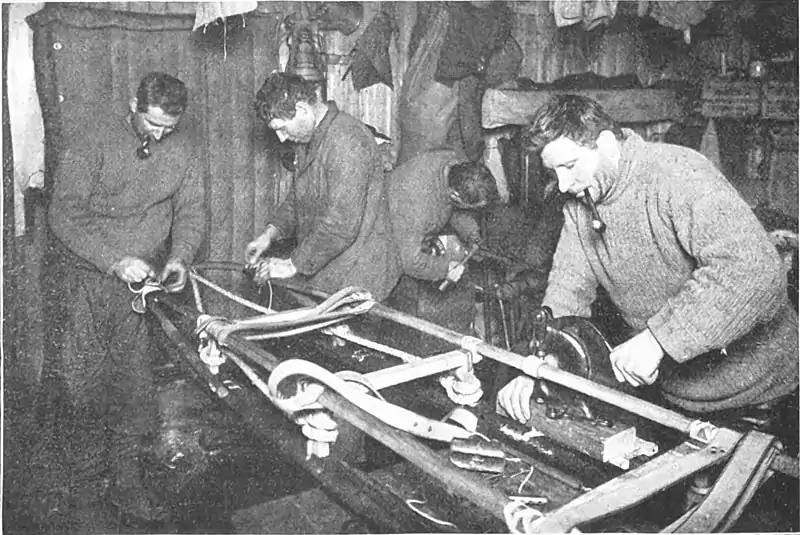 Photo of three men working on a sledge frame inside a hut, with a fourth in the background