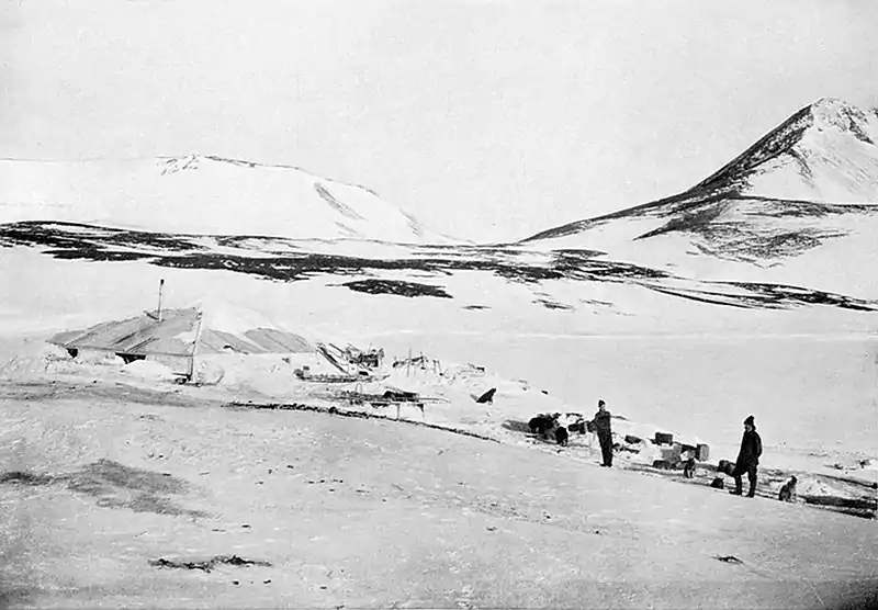 Photograph of two men in the middle distance standing amid supplies outside a low, snow-covered hut